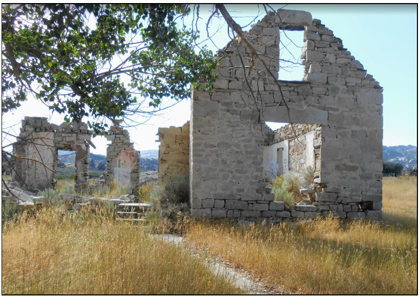 The crumbling stone walls and window frames of a roofless historic homestead ruin in a dry grassy field.
