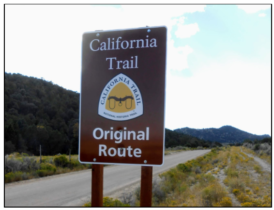 A dirt hiking trail winds through dry brush and green trees toward a large granite rock formation under a hazy sky.