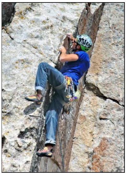 A rock climber wearing a helmet and blue shirt scales a steep vertical crack on a granite rock formation.