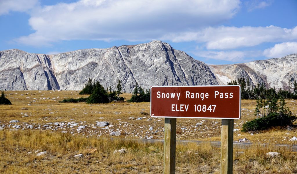 A brown wooden highway sign marking Snowy Range Pass Elev 10847 with the light-colored Medicine Bow Mountains rising in the background.