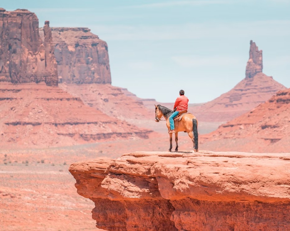 Man in red shirt riding a horse on a red rock overlook at John Ford’s Point in Monument Valley