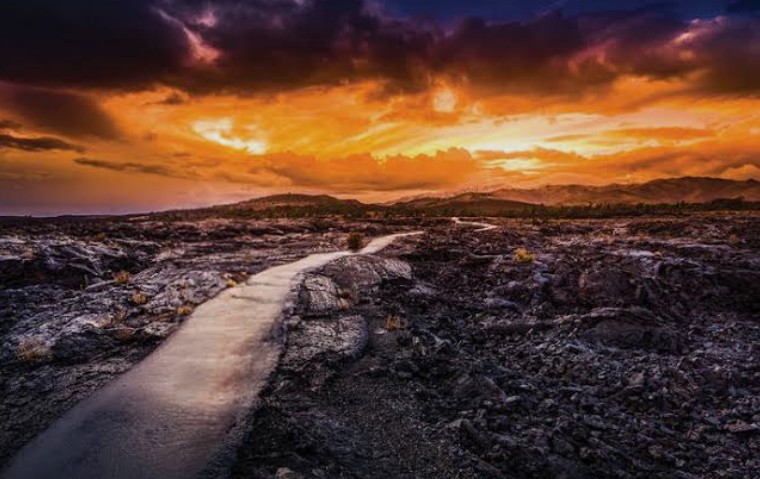 Panoramic view of a winding path through the black lava fields of Craters of the Moon National Monument under a dramatic orange and purple sunset.