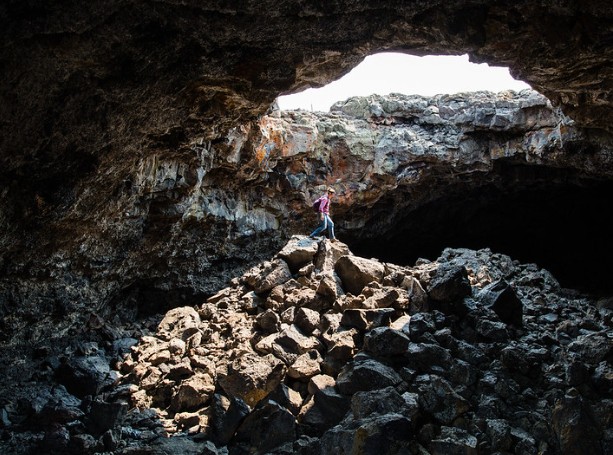 Interior view of a dark lava tube cave at Craters of the Moon with sunlight streaming through a collapsed ceiling onto volcanic rocks.