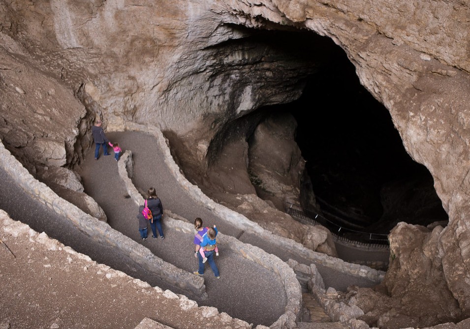 Visitors walking down a steep, winding paved switchback trail into a massive, dark cave opening.