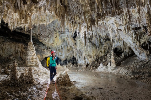 A hiker wearing a bright blue and yellow jacket stands on a cave trail surrounded by towering stalactites and columns.