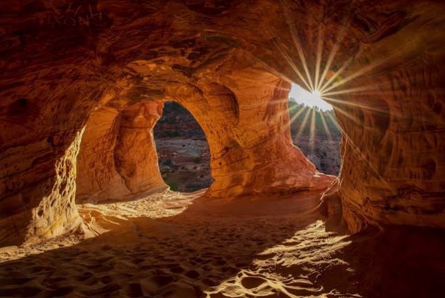 A dramatic sunburst shines through a natural rock archway inside a cave during sunset, casting warm golden light.
