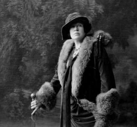 Vintage 1920s portrait of Margaret Molly Brown wearing a dark coat with large fur collar and cuffs, a matching cloche hat, and holding a walking cane.
