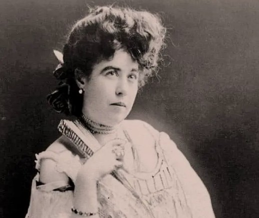 Vintage studio portrait of a young Margaret Molly Brown with an elegant updo hairstyle, wearing a Victorian lace gown, chokers, and a pearl earring.
