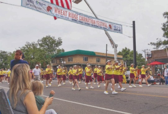 Parade marchers walking down 38th Avenue under the Wheat Ridge Carnation Festival banner.