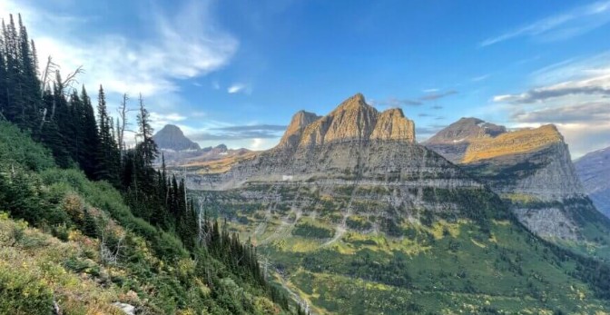 A wide panoramic view of rugged, sunlit mountain peaks towering over a green valley under a blue sky, with evergreen trees lining a steep hillside in the foreground.
