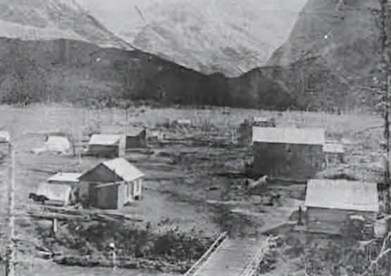 A historic black-and-white photograph from 1902 showing a small cluster of wooden cabins and dirt roads in a mountain valley.