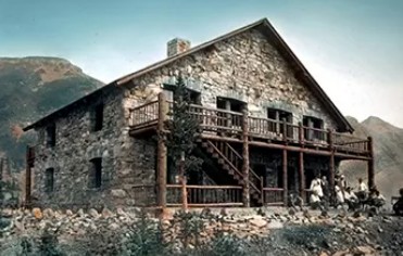 A historic colorized photograph from 1915 of a large, two-story rustic chalet built from stone and wood, featuring a prominent wooden balcony and stairs.