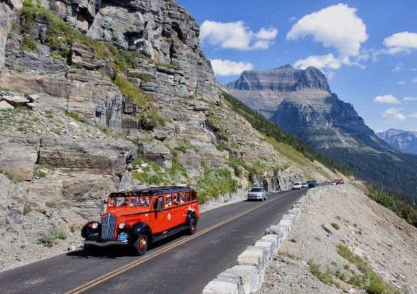 A classic red vintage tour bus driving along a narrow asphalt highway carved directly into the side of a massive, rocky cliffside under a bright blue sky.