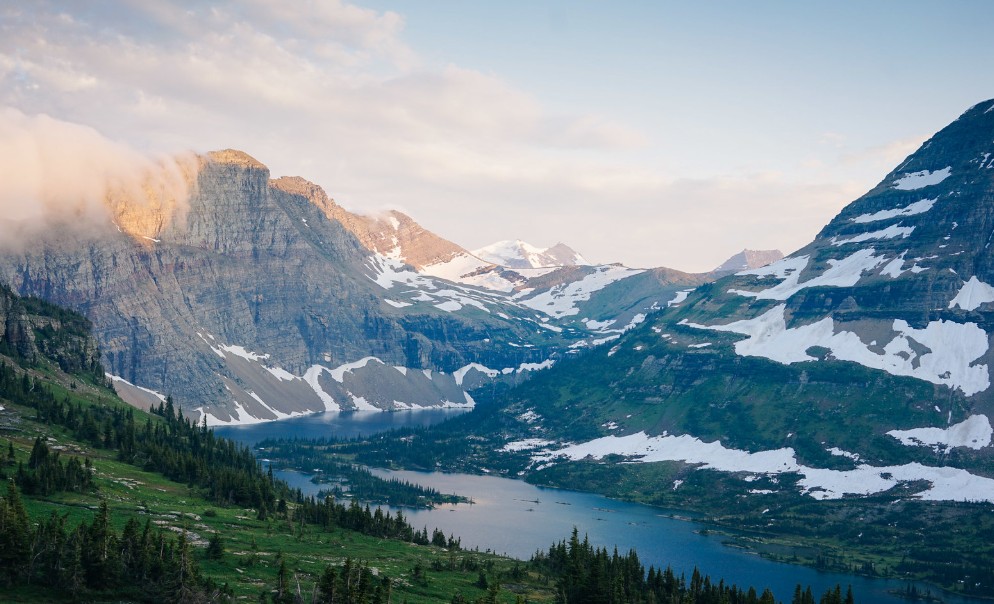 A scenic view of Hidden Lake nestled in a deep valley, surrounded by green forests and steep mountain slopes covered in patches of white snow and alpine ice.