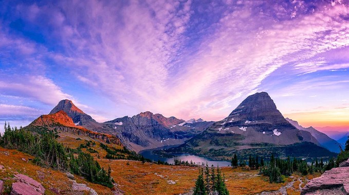 A breathtaking sunset view over Glacier National Park, featuring a vibrant purple and pink sky radiating over twin mountain peaks, alpine meadows, and a distant lake.