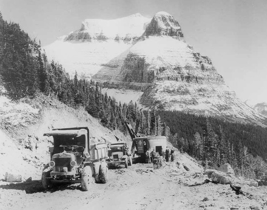 This historic photograph shows construction equipment, including trucks and a gas-powered shovel, working on the Going-to-the-Sun Road in Glacier National Park around 1932.