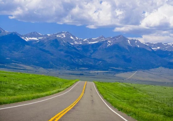 Two-lane highway winding through green fields toward Colorado mountain range.