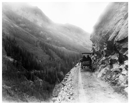 Black and white photo of an early model car on a narrow, dirt mountain cliff road in Colorado.