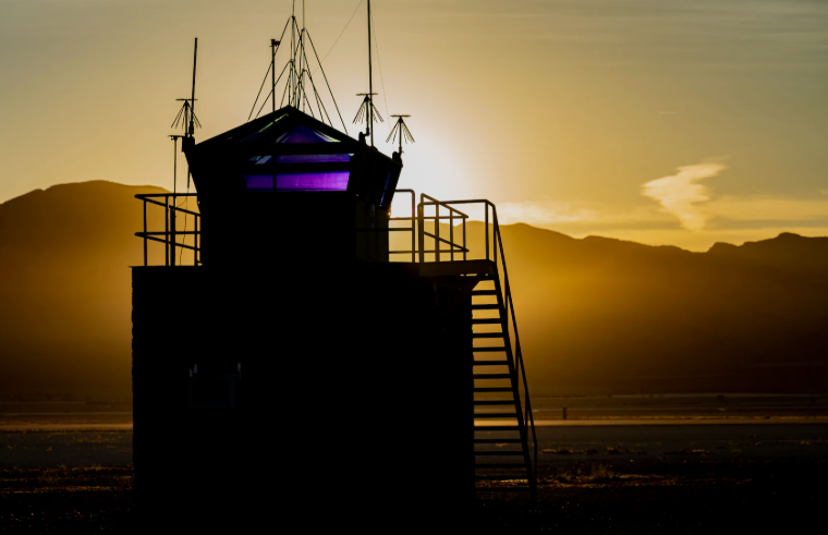 Silhouetted military control tower with glowing glass cab against a golden Nevada desert sunset.