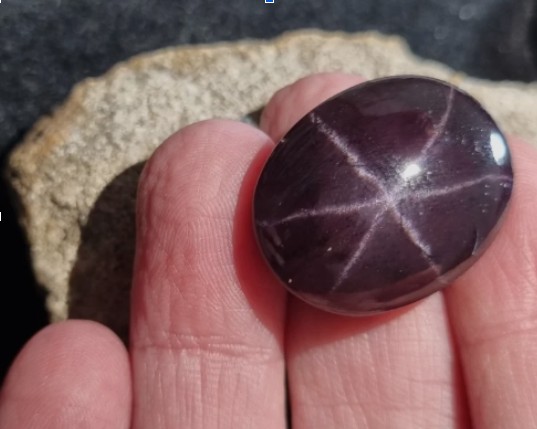 Oval cabochon six-rayed Idaho star garnet held between fingers over raw host rock.